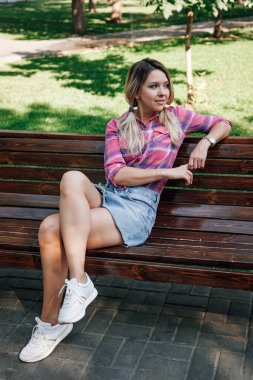 Yüzü dönük yetişkin sarışın bir kadın parkta bankta oturuyor, sol bileği sırtüstü yatmış, gündüzleri kareli gömlek ve kot etek giyiyor. Yeşillerle dolu sevimli bir arka plan