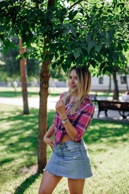 Yüzü kameraya bakan, yüzü parlayan sarışın genç bir kadın, bir eli cepte, diğeri ise parkta kareli gömlek ve etek giyerek duran buklelere dokunuyor. Yeşillerle dolu sevimli bir arka plan