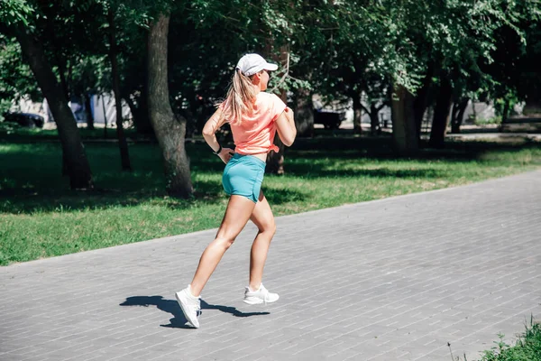 Bieganie młoda sportowa kobieta w parku w odzieży sportowej na tle zielonych drzew na szarych płytkach w słoneczny wietrzny letni dzień od tyłu — Zdjęcie stockowe