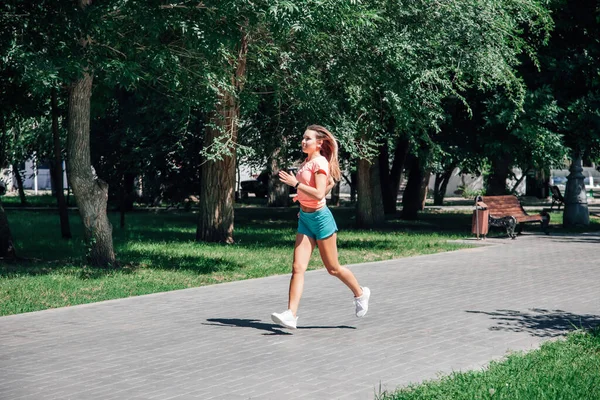 Running smiling young sporty woman in park in sportswear on background of green trees on grey tiles in sunny windy summer day — Stock Photo, Image