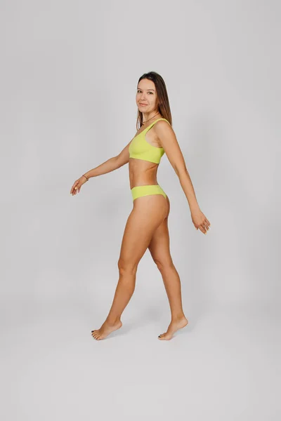 Morena posa sobre un fondo aislado. Mujer joven caucásica sobre fondo claro, caminando en ropa interior o traje de baño amarillo elegante, mostrando cuerpo delgado, tonificado perfecto, vista lateral — Foto de Stock