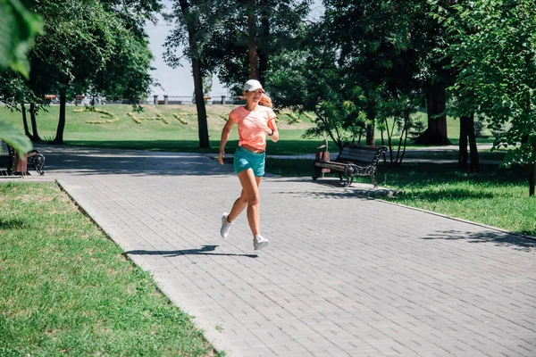 Correr sonriente joven mujer deportiva en el parque en ropa deportiva frente a los árboles verdes en la carretera de baldosas grises en el día de verano soleado —  Fotos de Stock
