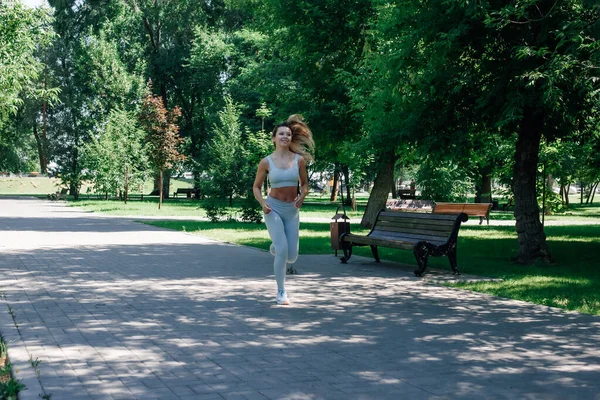 Biegnąca szybko uśmiechnięta młoda sportowa kobieta rozgrzewająca się, rozciągająca się w szarej odzieży sportowej w parku w słoneczny letni dzień — Zdjęcie stockowe