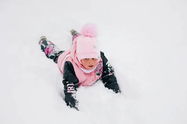 Χειμώνα πορτρέτο του γυναικείου παιδιού περπάτημα στη φύση κάνει άγγελος χιόνι, χαμογελώντας και ξαπλωμένος έξω στο χιόνι σε ζεστά ρούχα του χειμώνα με ξηρό γρασίδι γύρω. Αξιολάτρευτο φόντο γεμάτο χιόνι — Φωτογραφία Αρχείου