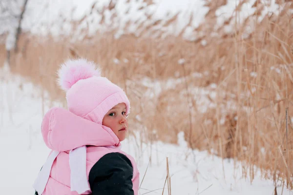 Closeup photo of female Russian kid with back to camera sitting in snow and looking behind wearing pink winter clothes in forest. Astonishing background full of white color and snow — Stockfoto