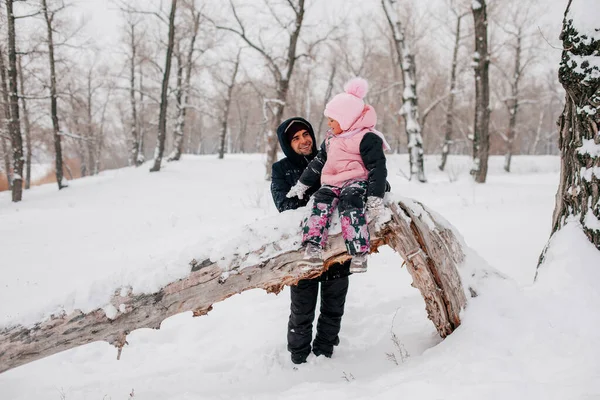 Distant photo of female kid sitting on tree log wearing pink winter clothes looking at smiling father standing behind daughter in forest. Astonishing background full of white color and snow — Stockfoto