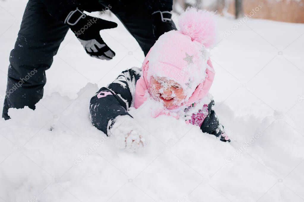  kid lying in snowdrift wearing pink winter clothes and father helping daughter to stand up in forest. Astonishing background full of white color and snow 