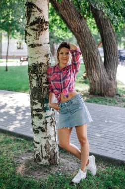 Yüzü kameraya bakan yetişkin sarışın kadın parkta dikiliyor huş ağacına yaslanıyor kareli gömlek giyiyor göbeğine bağlı ve kot etek giyiyor. Yeşillerle dolu sevimli bir arka plan