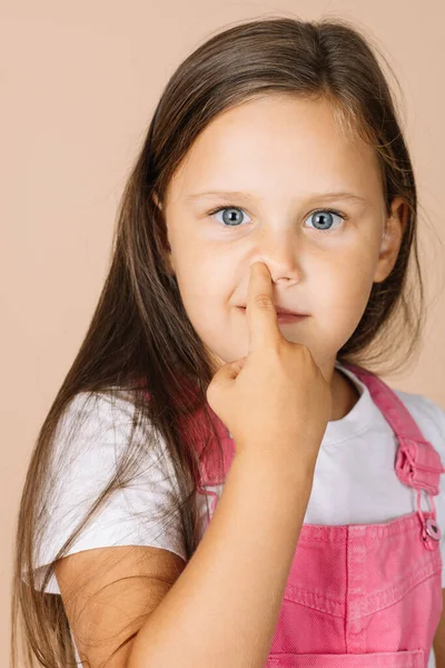 Close-up portrait female kid picking nose with point finger with shining eyes smiling looking at camera wearing bright pink jumpsuit and white t-shirt on beige background — Stock Photo, Image