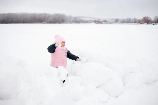 Closeup photo of female kid who is about to throw snowball at someone and smiling wearing pink winter clothes in park. Astonishing background full of white color and snow — Stockfoto