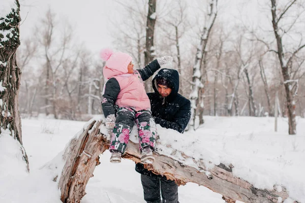 Distant photo of female kid sitting on tree log wearing pink winter clothes throwing snow at father standing behind daughter in forest. Astonishing background full of white color and snow — Stockfoto