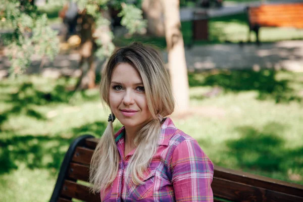 Portrait of adult blonde shining woman with face looking into camera sitting on bench in park wearing checkered shirt in daytime. Adorable background full of green — Stock Fotó