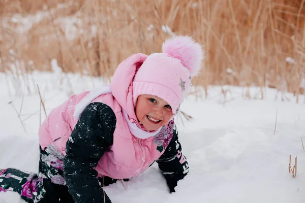 Крупним планом фотографія дівчинки, яка дивиться в камеру, плаче в снігу і посміхається зубами в рожевому зимовому одязі в лісі. Дивовижний фон, повний білого кольору і снігу — стокове фото
