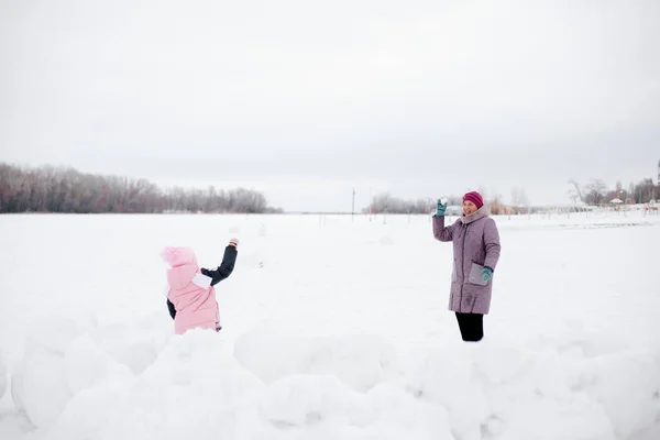 Enfant et grand-mère jetant des boules de neige à l'autre souriant s'amusant à porter des vêtements d'hiver roses. Fond étonnant plein de couleur blanche et de neige — Photo