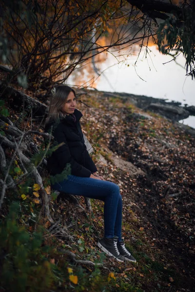 Świecąca młoda kobieta siedząca na korzeniach drzew w lesie, uśmiechnięta, odwracająca wzrok i pozująca obok rzeki z drzewem w tle, ubrana w niebieskie dżinsy, czarną kurtkę i szare trampki. Jesienny spacer w lesie — Zdjęcie stockowe