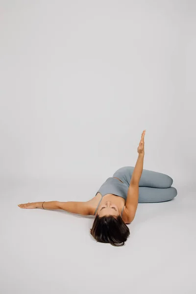 布鲁内特从事体育活动。年轻女子通过瑜伽或健身进行运动，她仰卧在床上，举起一只胳膊，双腿屈膝，灰色孤立的背景，后视镜 — 图库照片
