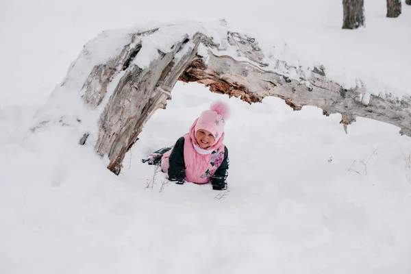 Distant photo of female kid looking into camera lying under tree log on snow and smiling wearing pink winter clothes in forest. Astonishing background full of white color and snow — Stockfoto
