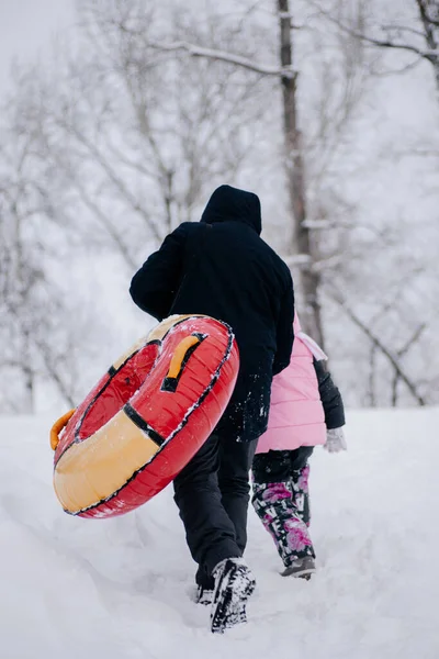 Дитина позаду батька з санчатами піднімається на пагорб, щоб ковзати в снігу в лісі. Дивовижний фон, повний білого кольору і снігу — стокове фото