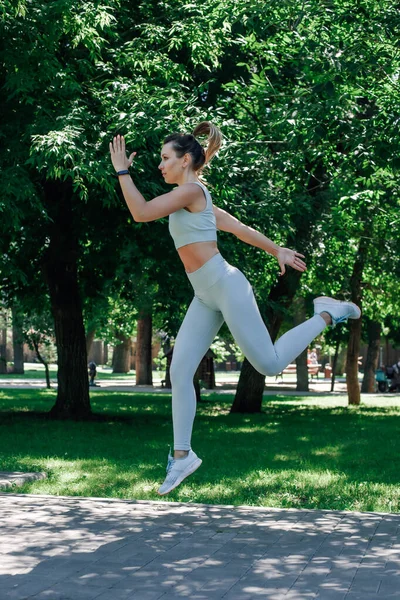 Attractive young sporty woman jumping in air stretching out arms joyfully in grey sportswear in summer sunny day in park — Stock Photo, Image