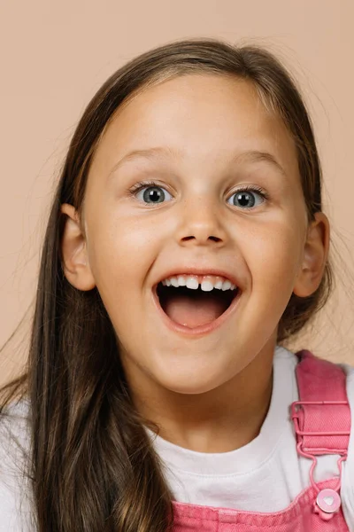 Niña con ojos brillantes y sonrisa excitada con dientes, boca abierta y cejas levantadas mirando a la cámara con mono rosa brillante y camiseta blanca sobre fondo beige — Foto de Stock