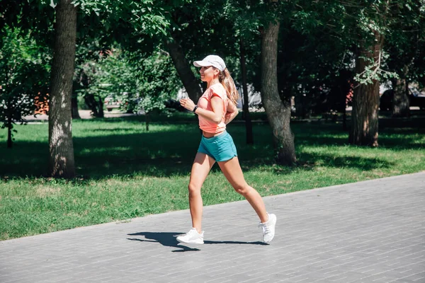 Corriendo sonriente joven mujer deportiva en el parque en ropa deportiva en el fondo de árboles verdes en baldosas grises en el soleado día de verano ventoso desde el frente —  Fotos de Stock