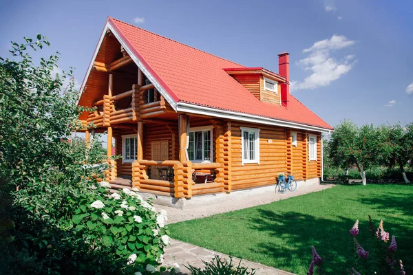 Kırmızı kiremit çatılı yeni yapılmış büyük ahşap bir kır evi, sağda çimenlik alan ve evin arkasında kiraz ağaçları bahçesi ve ön planda çiçek bahçesi. Köy hayatı 