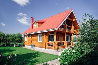 Kırmızı kiremit çatılı büyük ahşap kulübe evi, sol tarafta çimenlik alan ve evin arkasında kiraz ağaçları bahçesi ve ön planda da çiçek bahçesi. Köy hayatı 