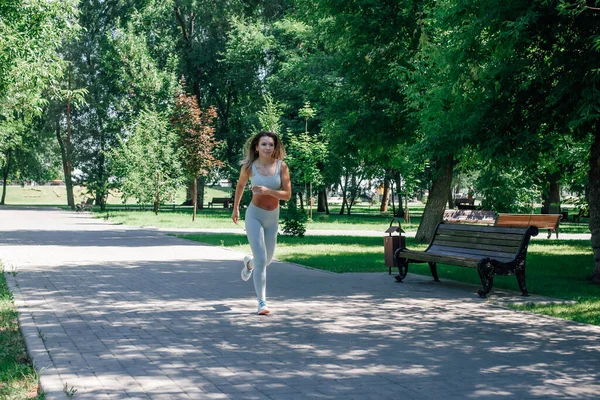 Running attractive young sporty woman warming up, stretching in grey sportswear in park in sunny summer day — Stock Photo, Image