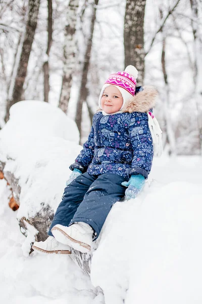 Κοριτσάκι που κάθεται καλυμμένο με χιόνι δέντρο κούτσουρο χαμογελώντας κοιτάζοντας κάμερα διασκεδάζοντας φορώντας ζεστά ρούχα του χειμώνα στο πάρκο. Φοβερό φόντο γεμάτο λευκό χρώμα — Φωτογραφία Αρχείου