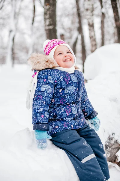 Щаслива молода дівчина сидить на дерев'яному колоді, вкритому снігом, дивлячись вгору і посміхаючись зубами в зимовому одязі в парку. Чудовий фон, повний білого кольору — стокове фото