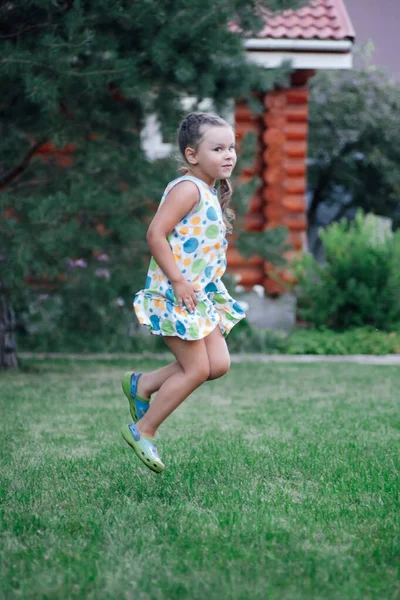 Jolie petite fille en saut souriant, regardant la caméra s'amuser en portant une robe d'été légère et des pantoufles dans le parc en journée. Fond incroyable plein de vert — Photo