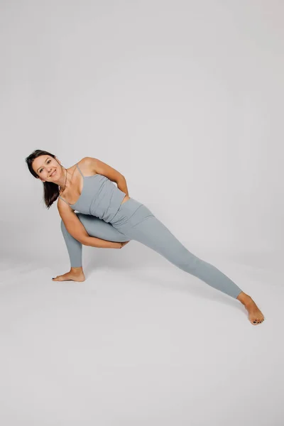 Sporty young woman does yoga. Concept of healthy lifestyle, brunette in gray fitness suit stands in yoga pose on isolated gray background, smiles and shows her flexibility — Stock Fotó