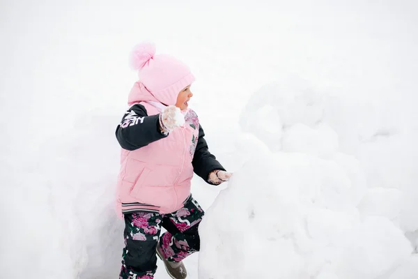 Closeup photo of female Russian kid who is about to throw snowball at someone and smiling in snowdrift wearing pink winter clothes in park. Astonishing background full of white color and snow — Stockfoto