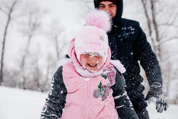 Closeup photo of female Russian kid who is about to cry because of snow in face wearing pink winter clothes and father standing behind in forest. Astonishing background full of white color and snow — стокове фото