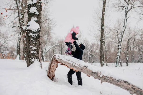 Bambino femmina in piedi sul tronco d'albero in procinto di saltare fuori indossando abiti invernali rosa con il padre che tiene figlia nella foresta. Sfondo sorprendente pieno di colore bianco e neve — Foto Stock