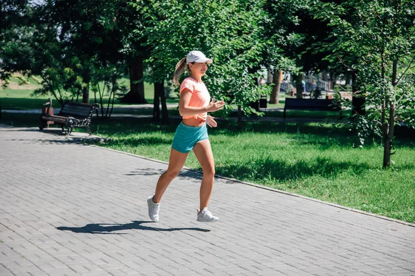 Running smiling young sporty woman in park in sportswear in front of green trees, lawn and bench on grey tiles in sunny summer day — Stock Photo, Image