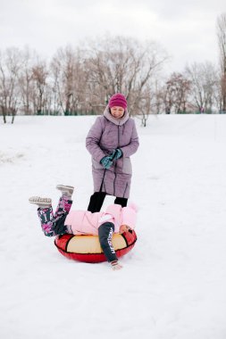 Kızaklı bir çocuğun uzaktan çekilmiş fotoğrafı. Büyükannesi torununu zahmetle çekiyor. Hem neşeli, hem de parkta pembe kışlık kıyafetler giyiyor. Beyaz renk ve karla dolu şaşırtıcı bir arka plan. 