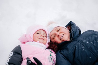 Annesiyle karda yatan ve ormanda pembe kışlık giysiler giymiş bir çocuğun yakın plan fotoğrafı. Beyaz renk ve karla dolu şaşırtıcı bir arka plan. 