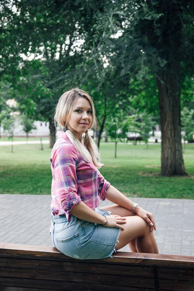Adult blonde womans side with face looking into camera sitting on bench in park with right hand on hip wearing checkered shirt and jeans skirt in daytime. Adorable background full of green — Stock Photo, Image