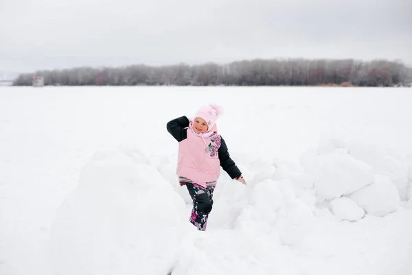 Gosse qui lance boule de neige à la caméra, souriant et regardant dans la caméra portant des vêtements d'hiver rose dans le parc. Fond étonnant plein de couleur blanche et de neige — Photo