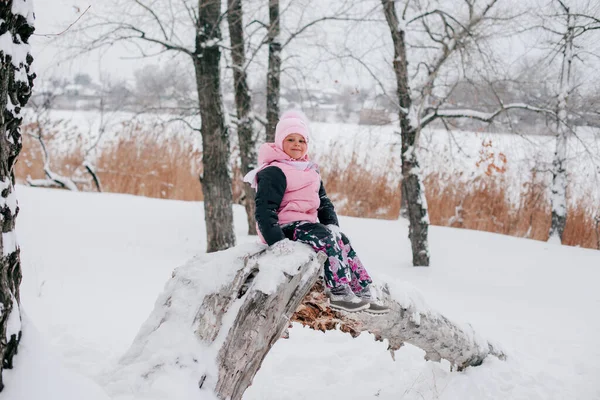 Distant portrait photo of female Russian kid looking into camera sitting on tree log and smiling wearing pink winter clothes in forest. Astonishing background full of white color and snow — Stockfoto