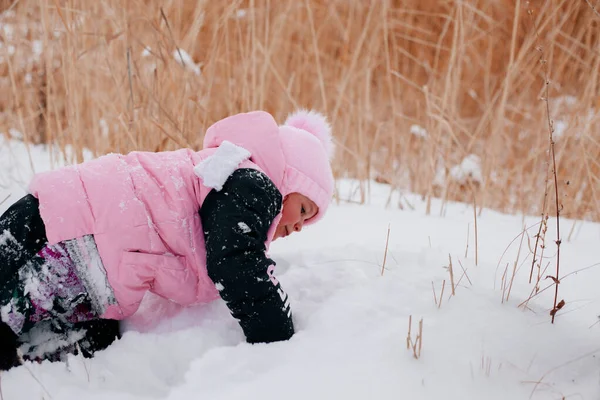 Foto di primo piano di una bambina russa che striscia nella neve e guarda la neve che indossa abiti invernali rosa nella foresta. Sfondo sorprendente pieno di colore bianco e neve — Foto Stock