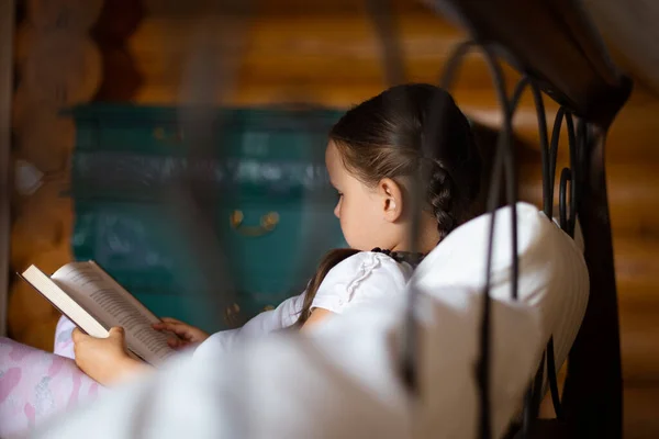 Молодая читающая девушка сторона захвачена из-за кровати лежал на подушке держа книгу в руках глядя на чтение книги с комодом на заднем плане в деревянном доме — стоковое фото