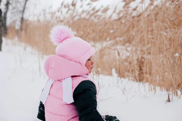 Foto di primo piano di una bambina russa con la schiena alla macchina fotografica seduta sulla neve e distogliendo lo sguardo indossando abiti invernali rosa nella foresta. Sfondo sorprendente pieno di colore bianco e neve — Foto Stock