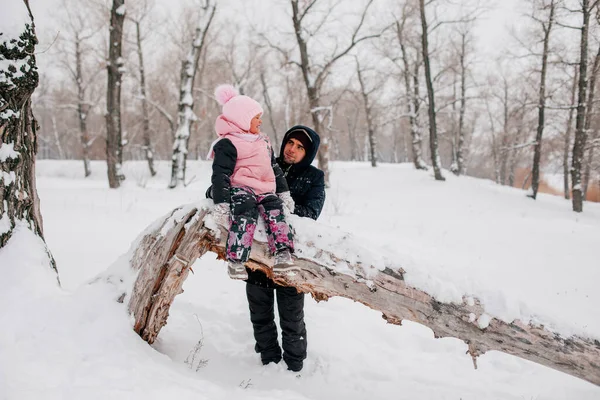 Distant photo of female kid sitting on tree log wearing pink winter clothes looking at loving father standing behind daughter in forest. Astonishing background full of white color and snow — Stockfoto