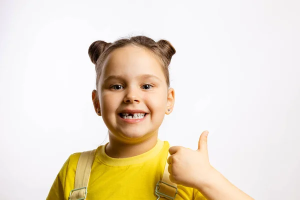 Retrato de una niña brillante que muestra un diente de bebé delantero faltante y el pulgar hacia arriba en una camiseta amarilla sobre fondo blanco. Los primeros dientes cambian. Ir al dentista para hacer el tratamiento dental — Foto de Stock