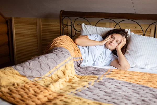 Sad curly brunette woman lying on bed under terry covers with worried face having inner problems wearing home pajamas in daytime. Anxiety moment in adult life — Zdjęcie stockowe