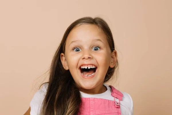 Cara de niño de cerca con ojos brillantes y sonrisa sobreexcitada con dientes, boca abierta y cejas levantadas mirando a la cámara con mono rosa brillante y camiseta blanca sobre fondo beige — Foto de Stock