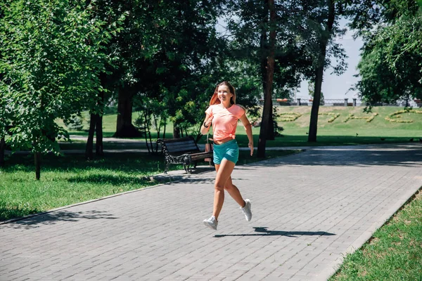 Running smiling young sporty woman in park in sportswear on background of green trees on grey tiles in sunny summer day — Stock Photo, Image