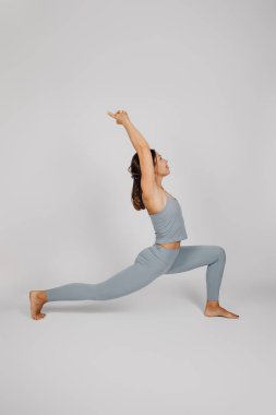 Genç bir kadın poz veriyor. Spor kıyafetli esmer, yoga yapıyor, savaşçı pozu veriyor. Gri arka plan, kenar görüş, sağlıklı yaşam tarzı konsepti.